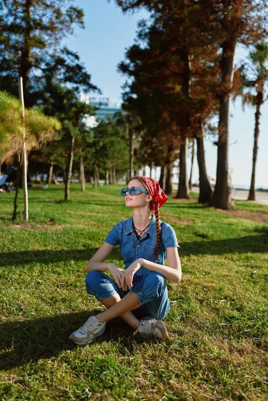 Lifestyle woman in modern boho-western denim street style sitting outdoors on grass with sunglasses and bandana, film color aesthetic, relaxed urban fashion vibe.