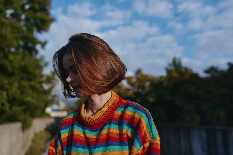 Young woman in a colorful striped sweater with bob haircut looks contemplative in an outdoor portrait on a suburban path during golden hour, casual outfit, relaxed mood.