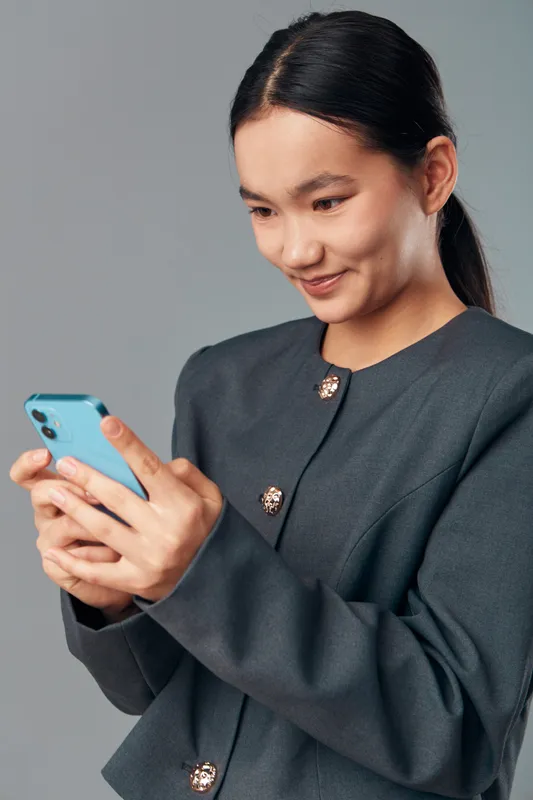 Young woman smiling and holding smartphone in hands looking at screen with interest wearing dark jacket on gray background. Closeup of female using mobile device for communication.