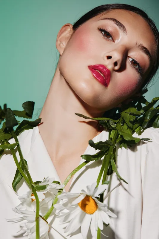 Portrait of young woman with red lipstick and lush green floral accents wearing white blouse on green background. Concept of beauty, fashion, and creative styling.