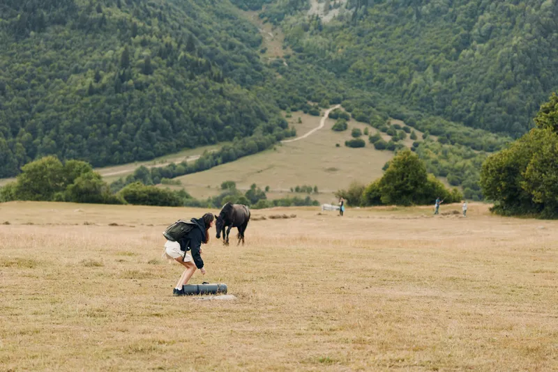 A solitary person in casual clothing tends a horse in a wide field beneath rolling hills. The calm scene evokes freedom, connection, and peaceful rural atmosphere.