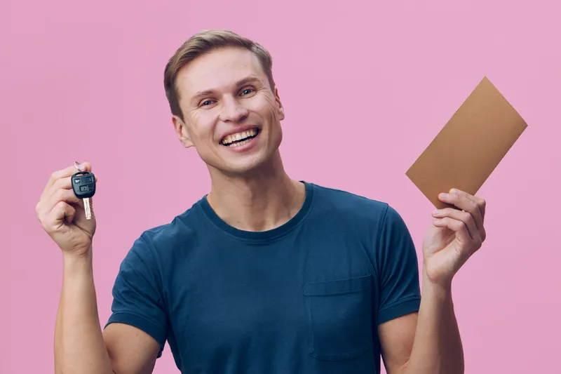 smiling young caucasian man hold car key document enjoy digital nomad lifestyle casual blue t-shirt pink background