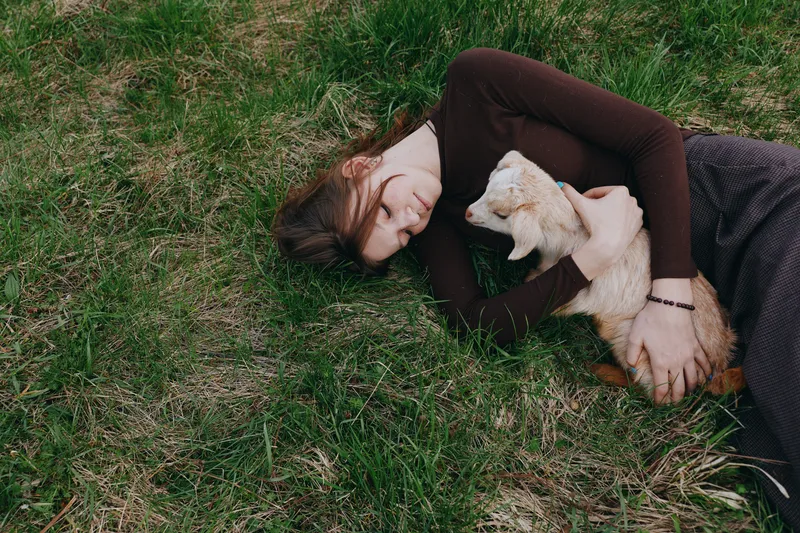 Young woman lies on grass, cuddling a light puppy, showing tender affection and a relaxed mood in an outdoor setting, peaceful companionship between person and pet in natural light.
