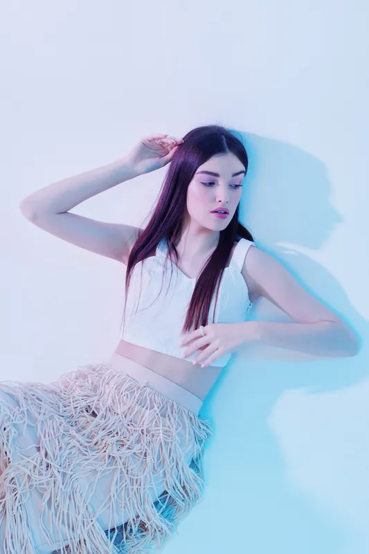 Young woman with long hair poses in a white crop top and fringed skirt. Soft blue and pink lighting highlights her beauty and fashion style in a studio setting.