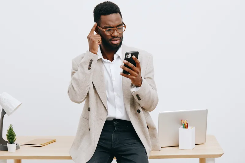 Professional man with glasses using smartphone while standing at workplace, dressed in smart casual in office setting, focused on screen, modern workspace interior, business communication concept.