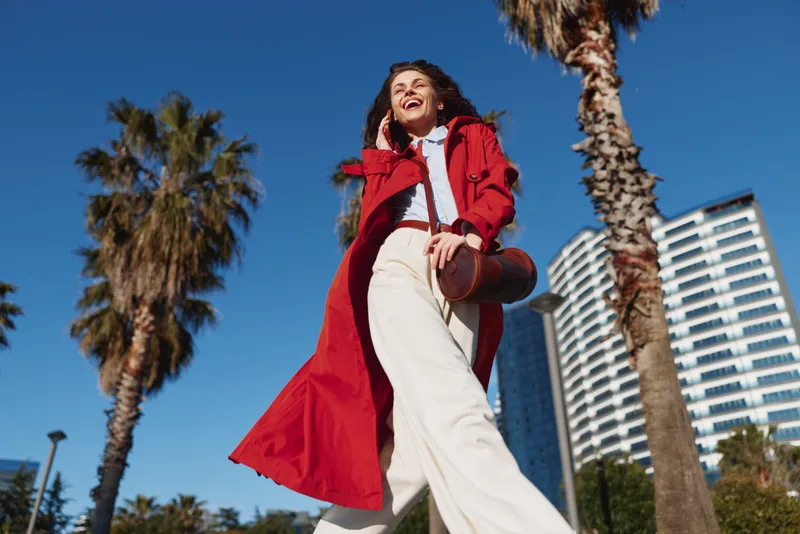 Happy woman in a red coat walks along a sunny city street with palm trees and modern buildings, showcasing energetic fashion and urban confidence under a clear blue sky.