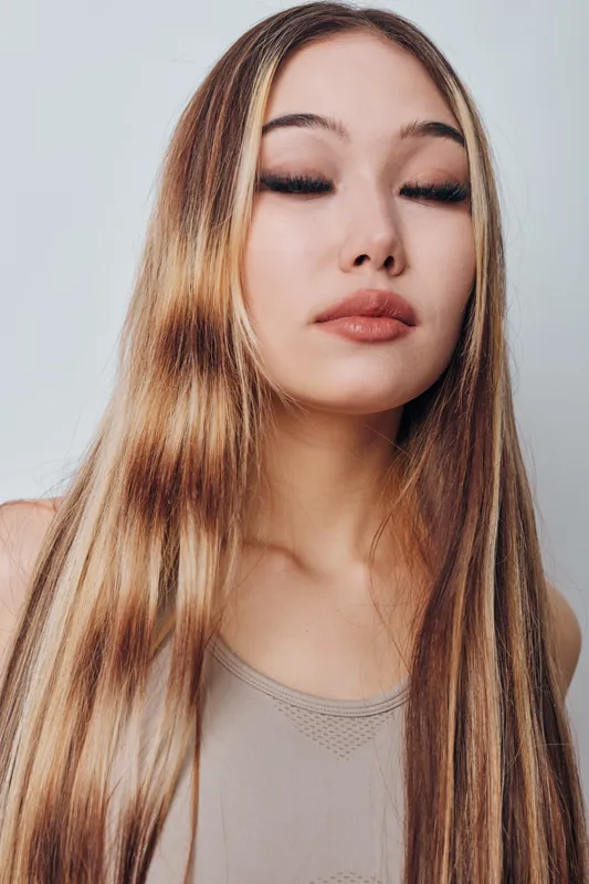 Young woman with long straight hair and closed eyes standing against plain background. Calm expression with natural makeup and relaxed posture indoors.