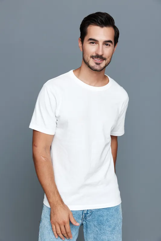 Smiling man in casual white t-shirt stands confidently in front of grey background, radiating positive energy and approachability. Lifestyle and fashion concept.