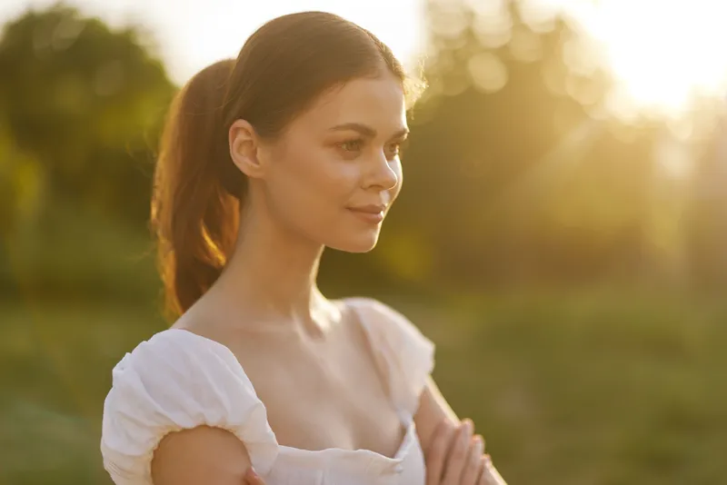 Young woman in a white dress enjoying a warm sunset, embodying serenity and beauty in a natural setting with soft sunlight glistening through the trees