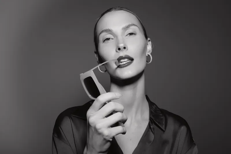 Elegant Caucasian woman with light skin holds sunglasses close to lips, wearing satin blouse and hoop earrings. Studio portrait in black and white, fashion and beauty concept.