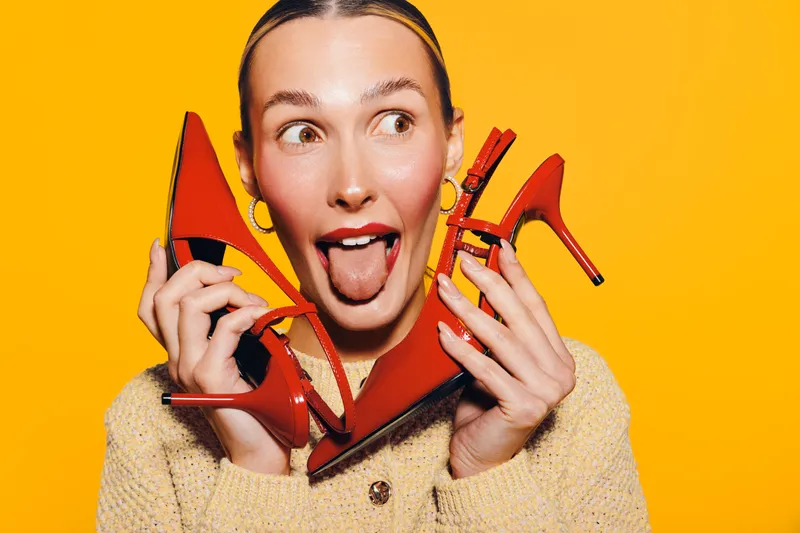 Young woman with slicked hair holds red high heel shoes near face, sticking out tongue. Bright yellow background creates vibrant mood. Concept for fashion, style, and playful attitude.