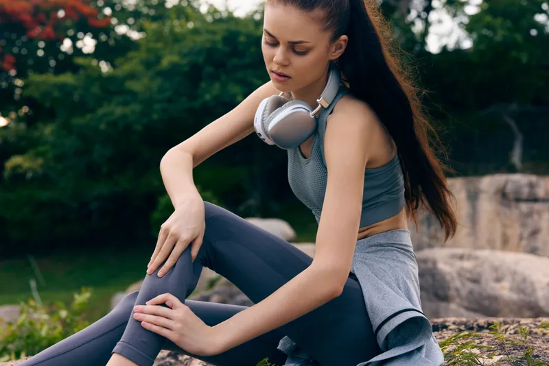 Young woman in stylish sportswear sitting outdoors, looking pensive, embodying fitness lifestyle and self care with headphones, nature backdrop