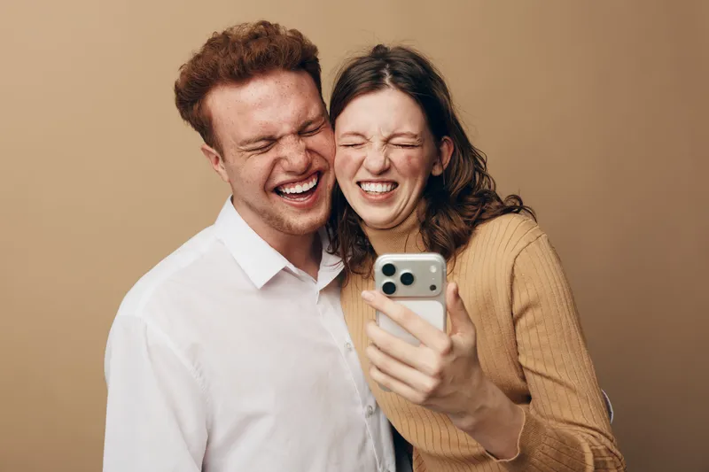 Happy young couple taking a selfie with smartphone, smiling and laughing together on beige background. Concept of friendship, joy, technology, and casual lifestyle.