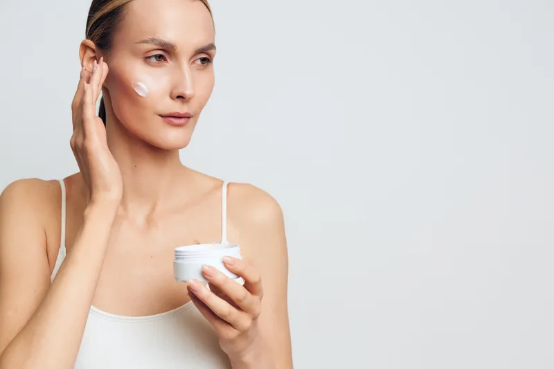 Young woman applies moisturizing cream on face holds jar with skincare product wears white tank top isolated on light background natural beauty care concept.