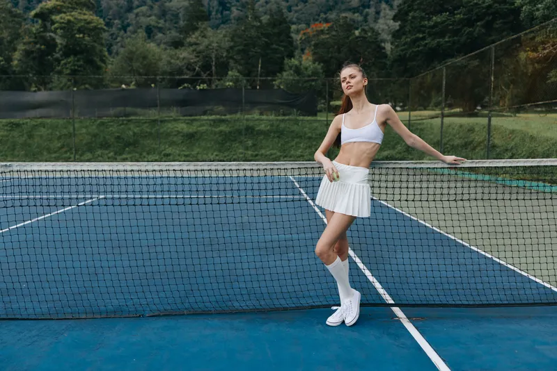 Young athletic woman in sporty outfit poses confidently on a tennis court, showcasing elegance and energy in her white sportswear and playful demeanor