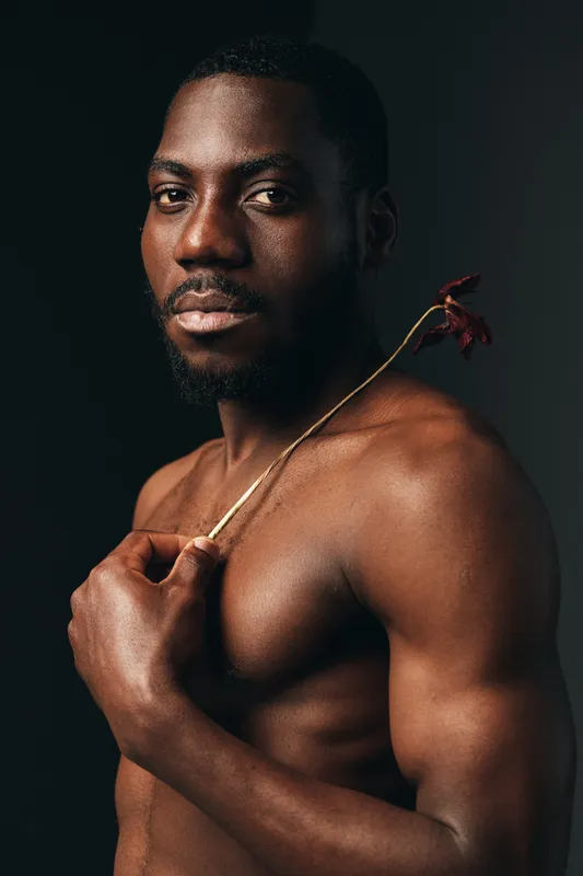 Black man with dark skin and muscular body holding dried flower on shoulder in creative visual design style with studio lighting and dark background.