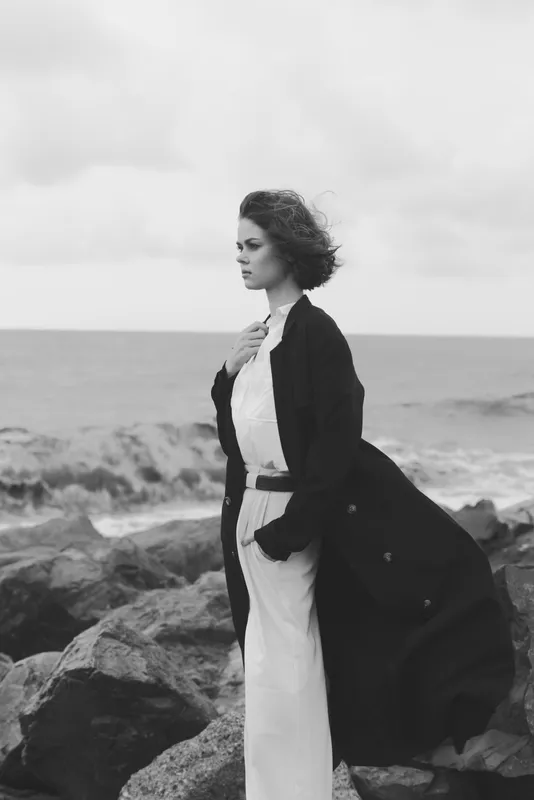 Woman on rugged rocks by the sea, wearing a long dark coat over a light dress, wind blowing hair, moody monochrome coastal fashion scene, ocean backdrop, thoughtful poised stance