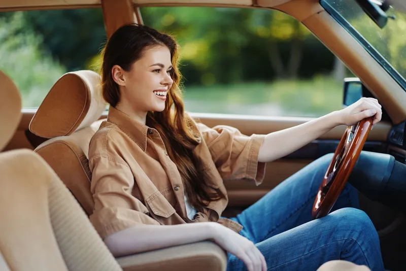 Young woman laughing inside a vintage car, wearing a casual beige shirt, enjoying a sunny day in nature, perfect for lifestyle and travel concepts.