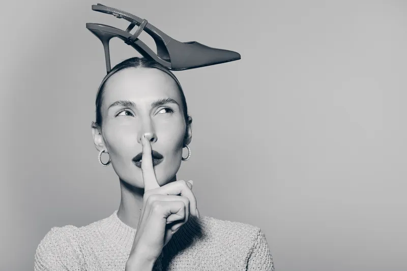 Young white woman with pale skin and dark hair holds finger to lips, balancing high heel shoe on head. Concept for silence, fashion, and creativity in studio.