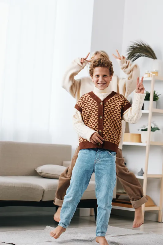 Joyful child dances indoors, wearing a warm knitted vest and cozy sweater, mid jump with arms raised, bright natural light, playful home setting and pure happy energy.