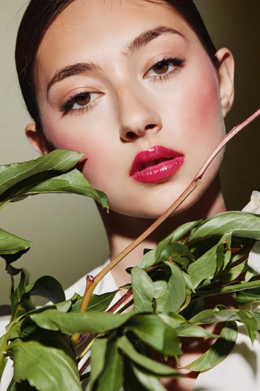 Fashion portrait of a young woman with glossy red lips and natural makeup holding lush green leaves on a plain background. Concept of beauty, elegance, and creative styling.