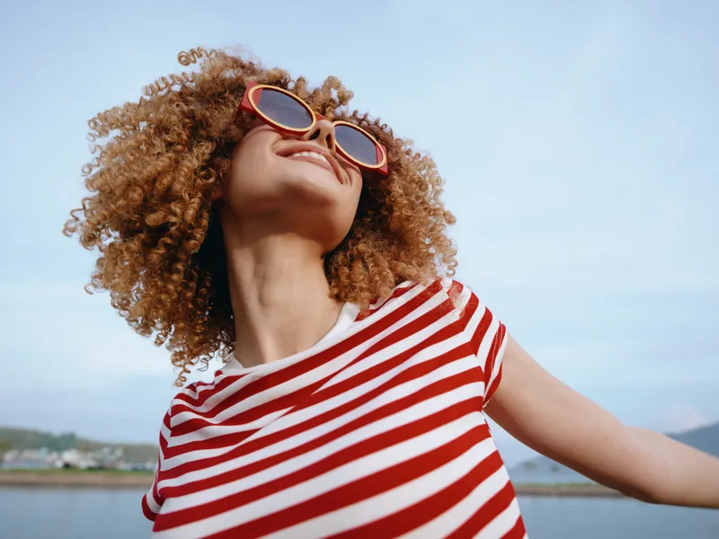 A young woman with curly hair wearing a red striped shirt and bold sunglasses stands outdoors, smiling toward the sky as a gentle breeze lifts her carefree expression.