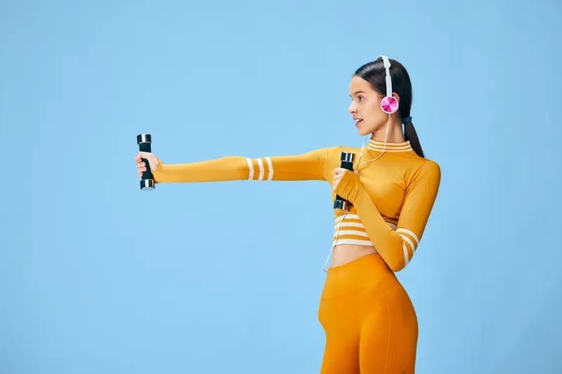 Young woman fitness portrait in orange activewear holding dumbbells and wearing pink headphones, performing arm exercise against a clean blue studio background.