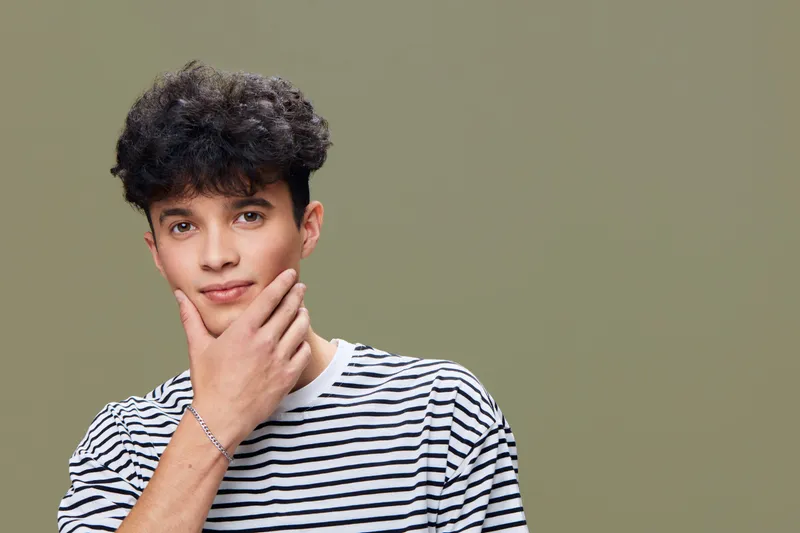 Young man with curly hair wearing a striped shirt posing thoughtfully against a muted green background, showing casual style and confident expression.
