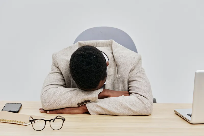 Videogame development concept image showing a young man with dark skin and curly hair resting his head on his crossed arms at a desk, surrounded by a laptop, smartphone, glasses, and notepad, suggesti