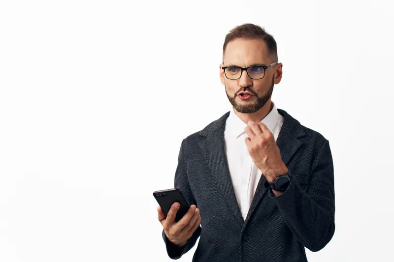 A confident business professional stands against a plain background, holding a smartphone and expressing focused emotion, thoughtful leadership, and readiness in a corporate portrait.