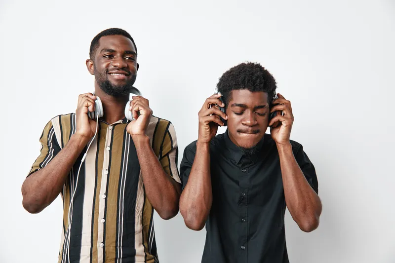 Two African American men with dark skin wearing casual shirts listening to music on headphones, smiling and enjoying sound in studio with white background.