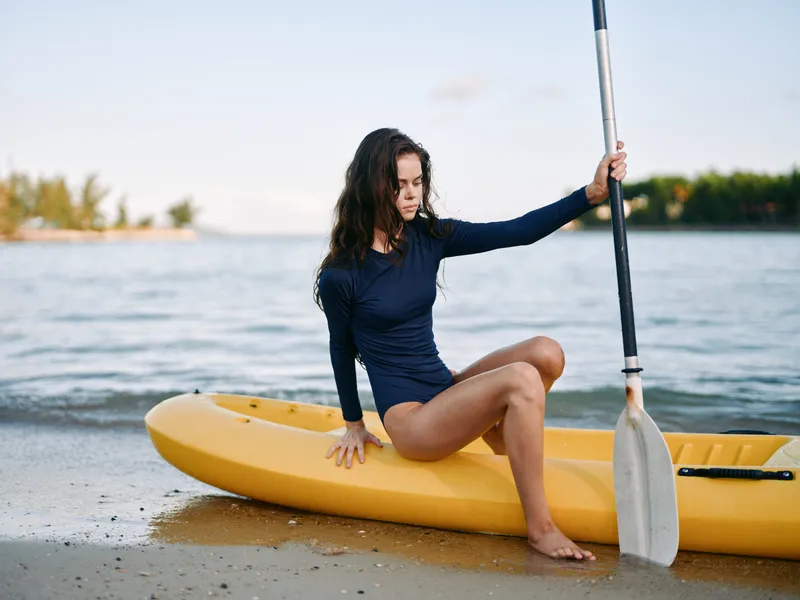 Young woman sits on a bright yellow kayak near the shoreline, wearing a navy long sleeve swimsuit, holding a paddle, gazing toward the calm sea with a relaxed expression.