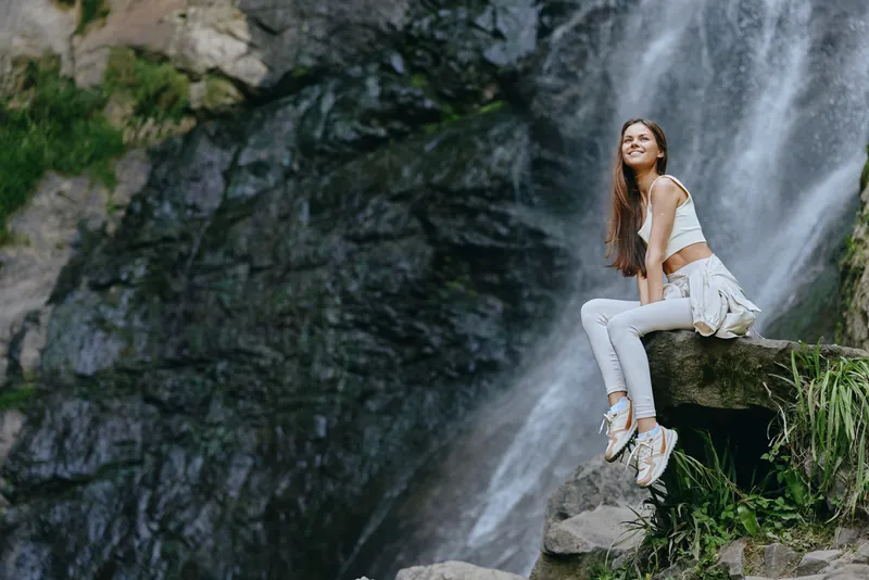 smiling woman sitting on rock near waterfall, wearing casual white outfit, enjoying nature, outdoor adventure, bright and natural light
