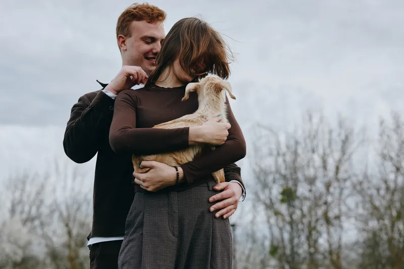 Cheerful couple outdoors embrace a playful goat kid, sunlit moment, warm tones, gentle breeze, natural textures, rustic countryside backdrop, affectionate interaction, lifestyle scene
