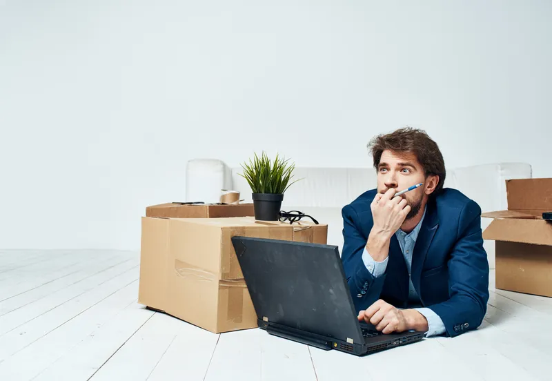 Мужчина в костюме лежит на полу среди офисных коробок, распаковывая ноутбук