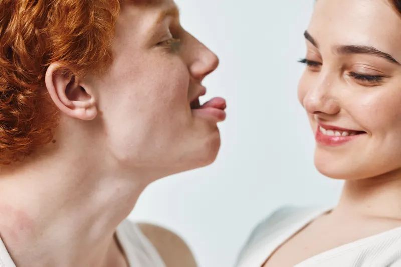 Young red-haired man playfully sticking out tongue at smiling woman with dark hair in casual studio portrait. Closeup, friendly interaction, light background, positive mood, youthful expression.