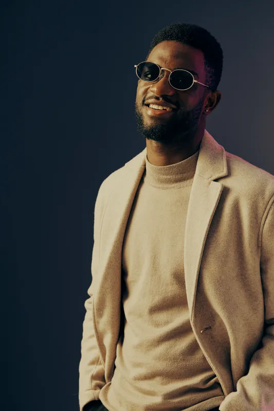 Stylish African American man in a beige coat and sweater smiling, showcasing modern fashion against a colorful studio background Seasonal autumn style portrait