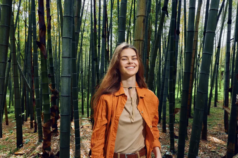 Woman smiling in bamboo forest nature portrait outdoors wearing orange jacket, happy young adult enjoying sunlight and green foliage, casual travel lifestyle image for wellness and joy.