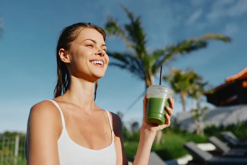 Young woman with wet hair enjoys a green smoothie outdoors on a sunny day, smiling and feeling refreshed in a casual white tank top near a poolside environment.