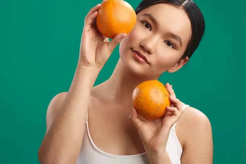 asian woman is pictured holding two oranges in a bright studio setting. she smiles softly, presenting a fresh, healthy lifestyle with a vibrant and relaxed mood.