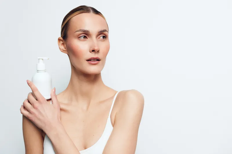 Young woman with clear skin holding a white lotion bottle on a light background. Concept of skincare, beauty routine, self-care, and natural health. Soft lighting highlights her smooth complexion.