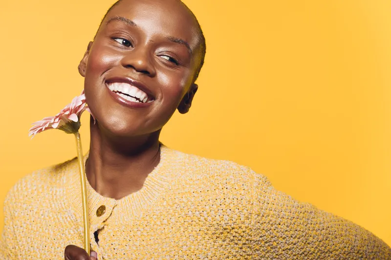 Happy African woman with dark skin smiles holding a flower on yellow background. Portrait of joyful female model in knitted sweater with natural beauty and positive mood concept.