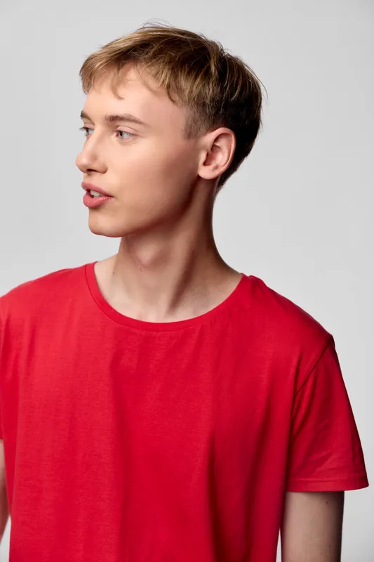 Young man in a red shirt speaks to the side in a clean studio setting, showcasing a candid conversational moment. Natural lighting, simple backdrop, versatile for lifestyle or communication themes.