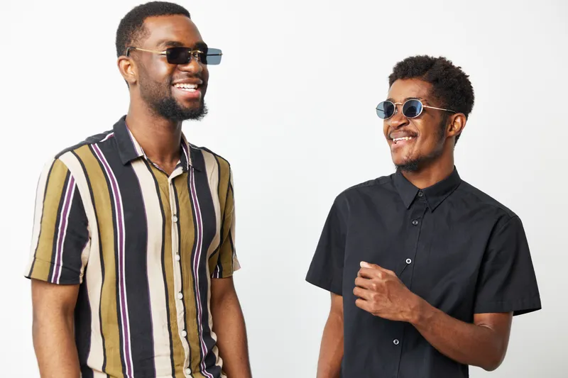 Two stylish African American men wearing sunglasses share a joyful moment together, showcasing their vibrant outfits and friendly smiles. Perfect for fashion or friendship themes.