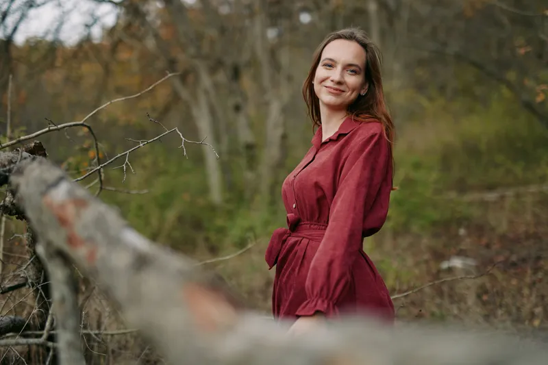 An authentic portrait of a woman in a burgundy dress posed beside a rustic fence, highlighting natural texture, color, and serene mood in a woodland setting with honest gleam.