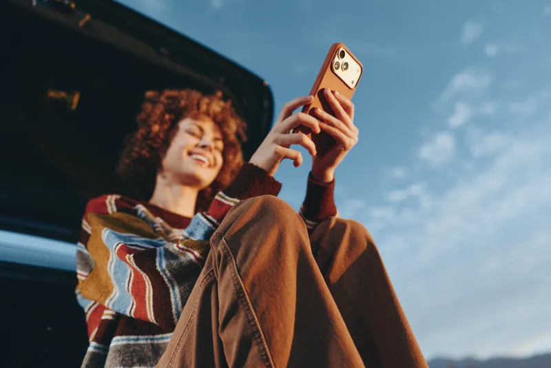 Woman lifestyle scene outdoors, smiling and holding smartphone, wearing winter coat, standing by car, bright sky and sunlight in background, relaxed tech moment for modern lifestyle
