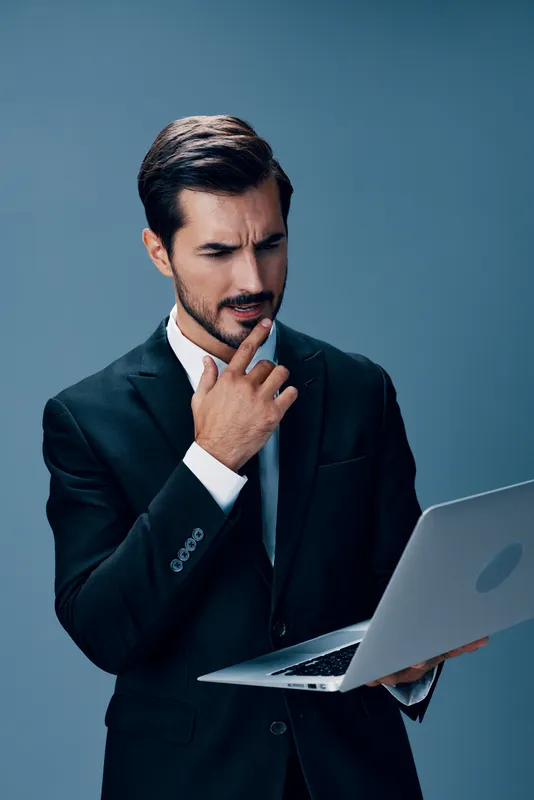 Уставший и испытывающий стресс мужчина в деловом костюме смотрит на ноутбук и работает онлайн через интернет во время видеозвонка и деловых переговоров на синем фоне
