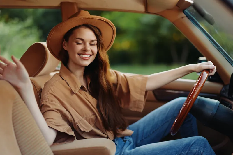 Young woman with long hair wearing a hat, smiling joyfully while sitting in a classic car, enjoying a sunny day outdoors.