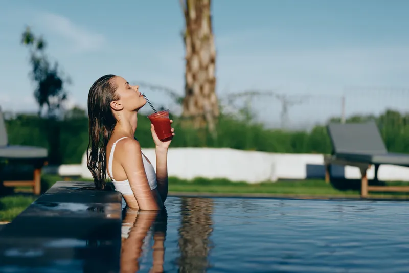 Smiling woman in a white swimsuit sips a red drink at the pool edge, water up to her shoulders. Bright outdoor setting conveys leisure, wellness, and carefree summer vibes.