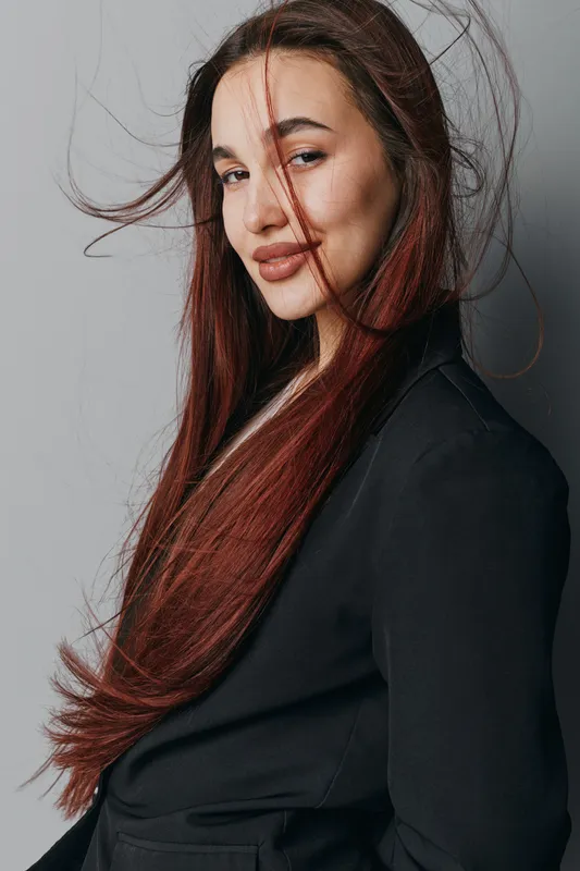 Woman with long red hair wearing black jacket posing with slight smile and wind blowing strands of hair in studio with gray background. Confident female model with natural makeup and casual style.
